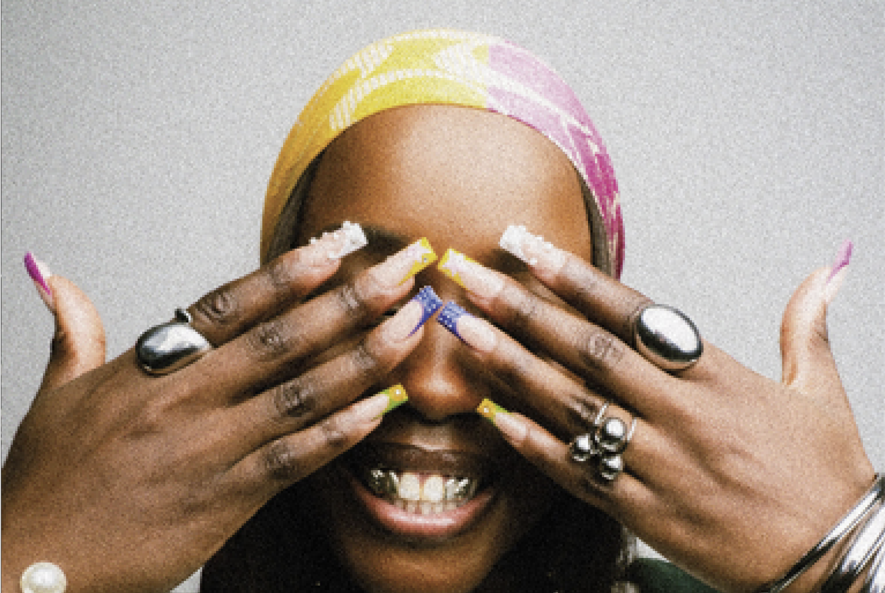 Person with colorful headscarf smiling, covering eyes with hands showing multicolored nails and silver rings.