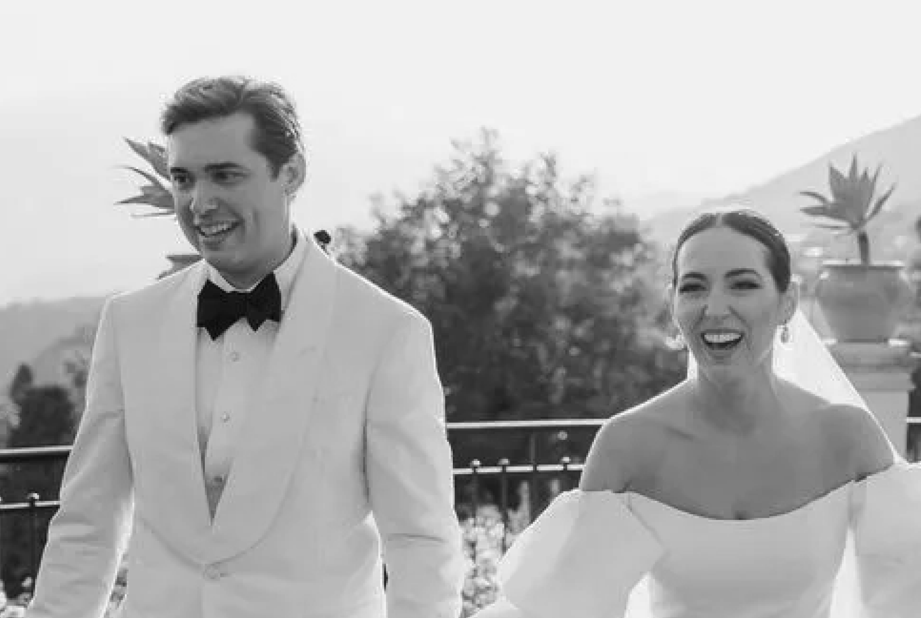 Smiling bride in off-shoulder dress and groom in white tuxedo with black bow tie outdoors.
