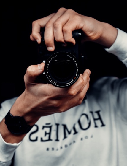 Person wearing a white long-sleeve shirt holding a camera facing the viewer against a black background.