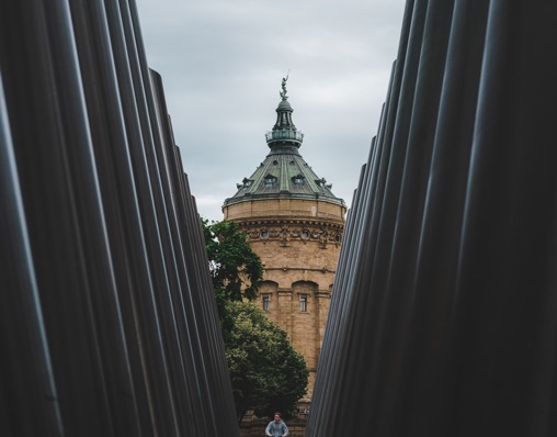 Historic tower viewed between large vertical dark pillars with trees and a person below on a cloudy day.