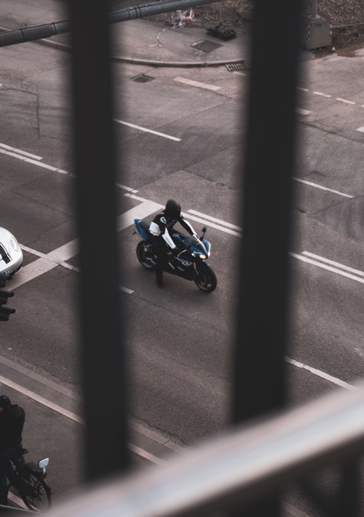 Person in black gear sitting on a blue motorcycle stopped at an intersection, viewed through black bars.