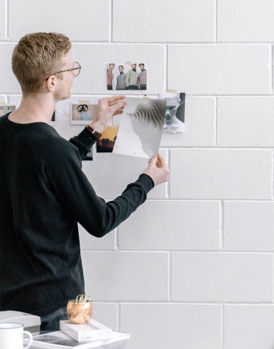 Man with glasses arranges photos on a white brick wall in a minimal workspace.