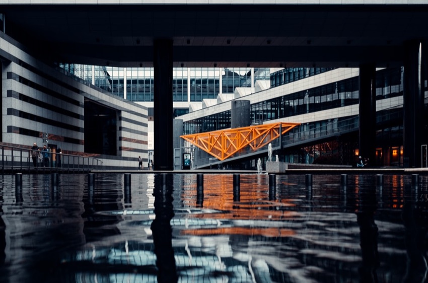 Modern building with an orange geometric structure reflected in calm water in the foreground.