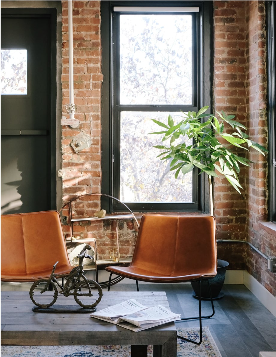 Two brown leather chairs beside a window with exposed brick walls and a potted plant.