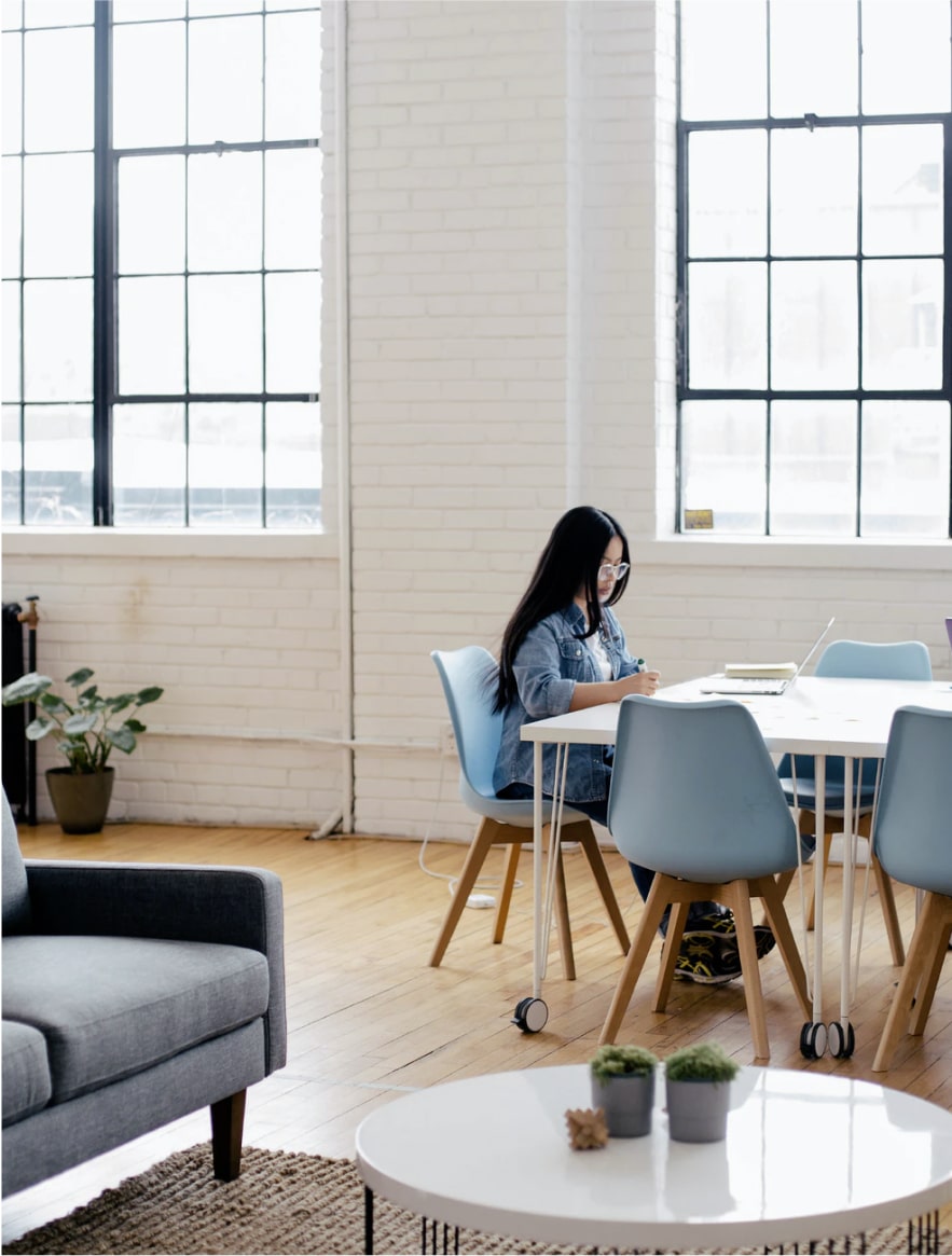 Woman with glasses writing at a table in a bright modern workspace with large windows.