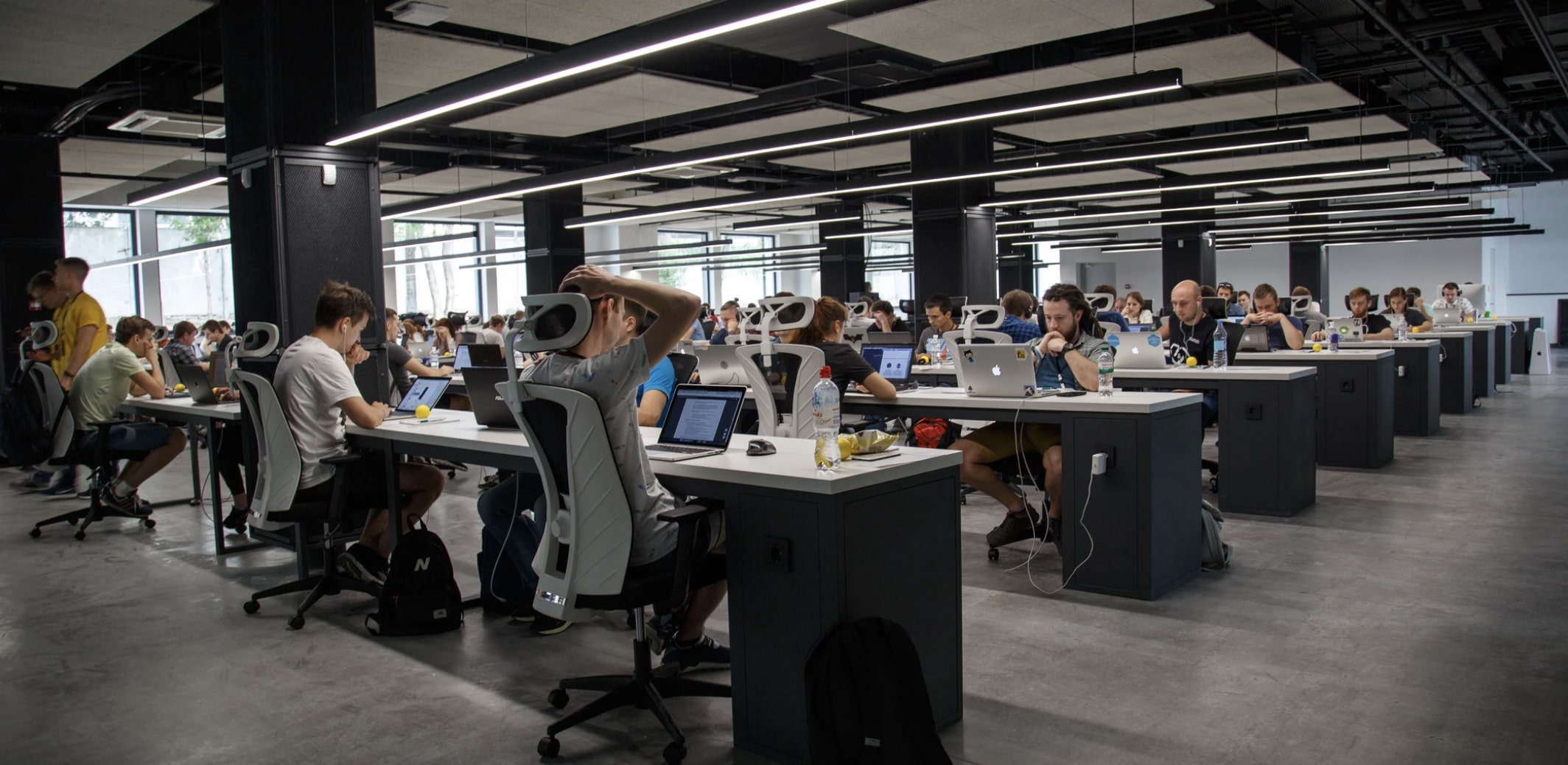 Modern open-plan office with rows of people working at desks with laptops and bottles.