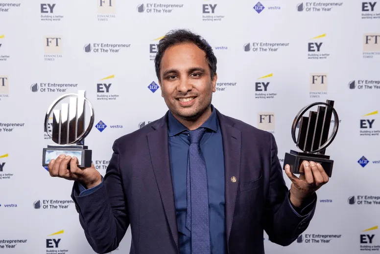 Man in suit and tie smiling, holding two EY Entrepreneur Of The Year awards in front of branded backdrop.