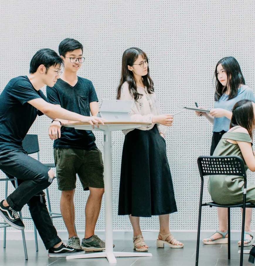 Group of young people standing and sitting around a table, discussing and sharing papers.