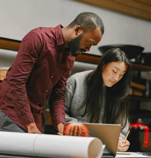 Man and woman working together, looking at a laptop and writing on paper at a desk.
