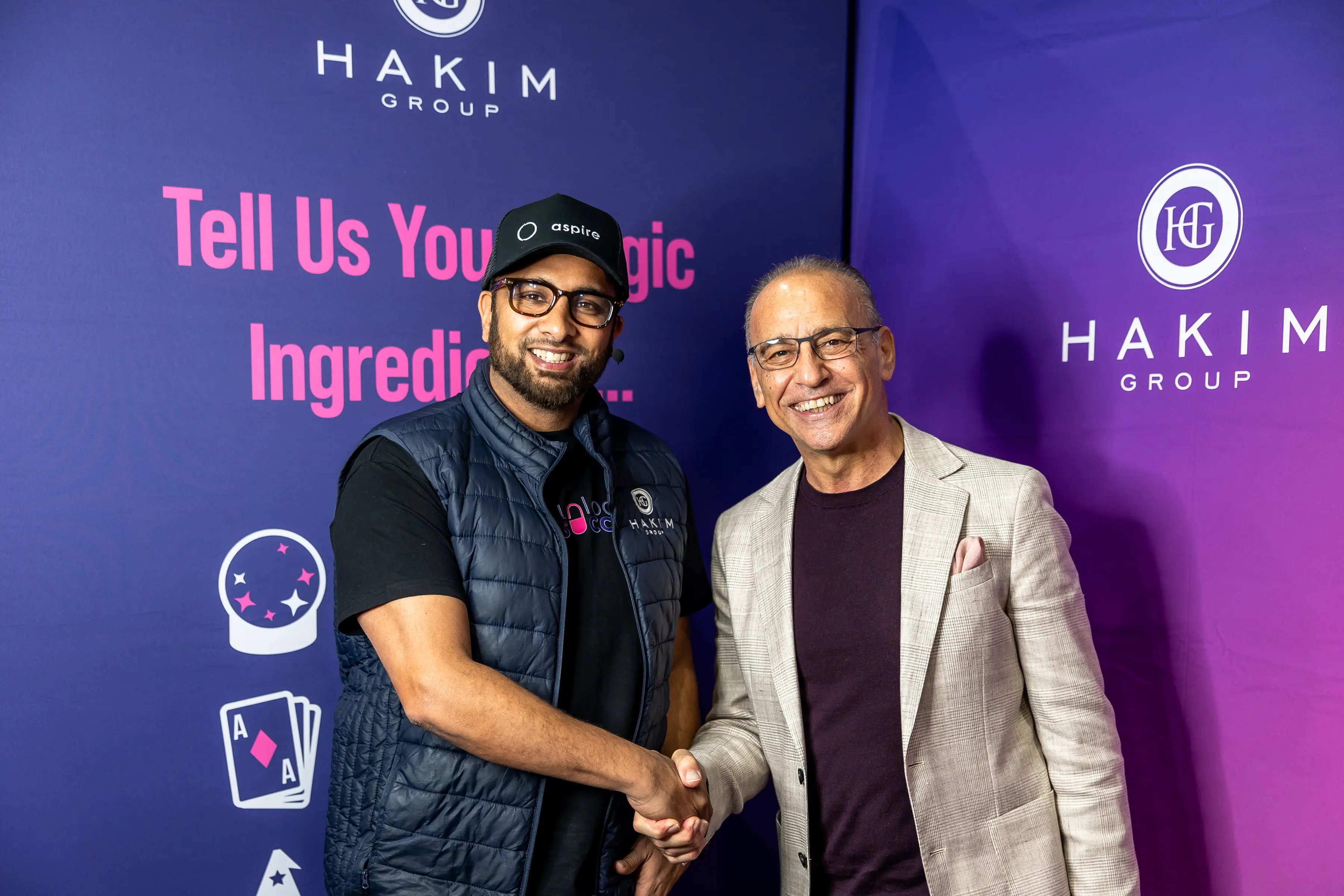 Two men smiling and shaking hands in front of a Hakim Group purple backdrop with text and logos.