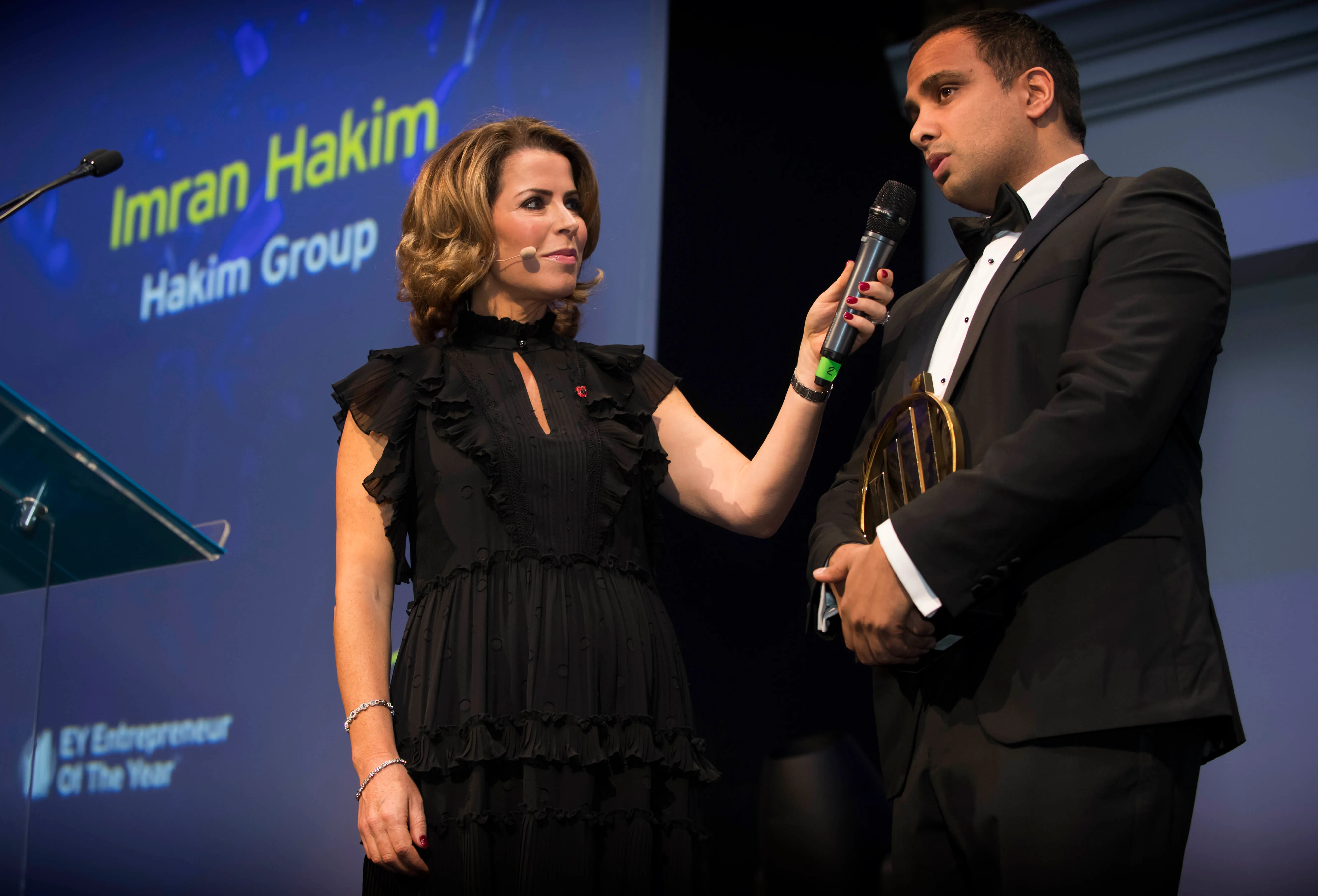 Man in tuxedo holding an award speaks into a microphone held by a woman in a black dress on stage.