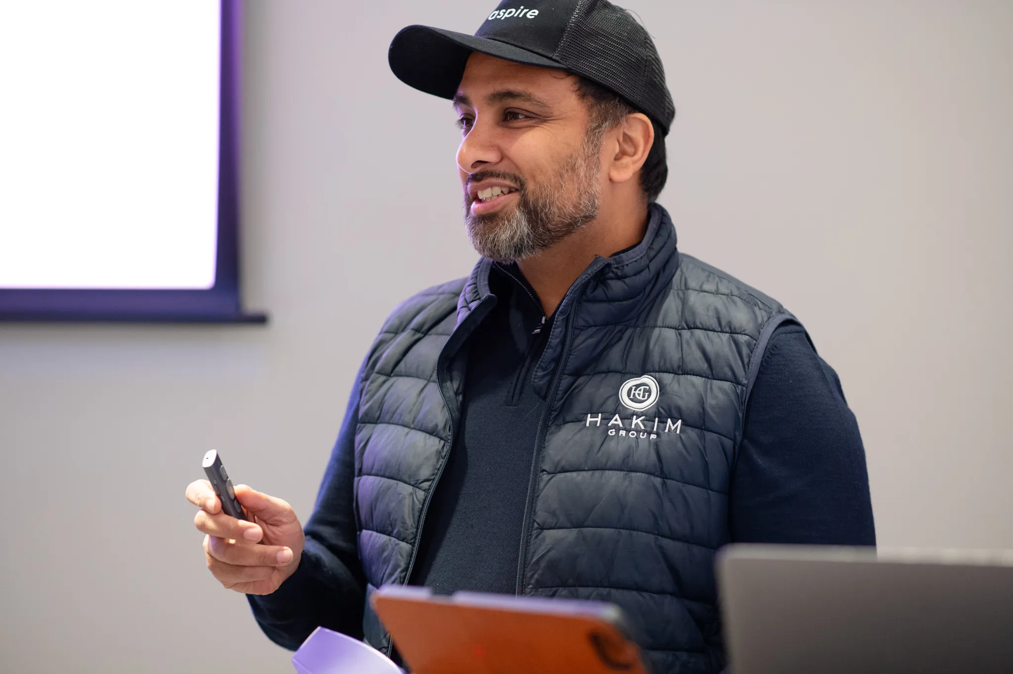 Man wearing a Hakim Group vest and Aspire cap, holding a presentation remote and speaking.