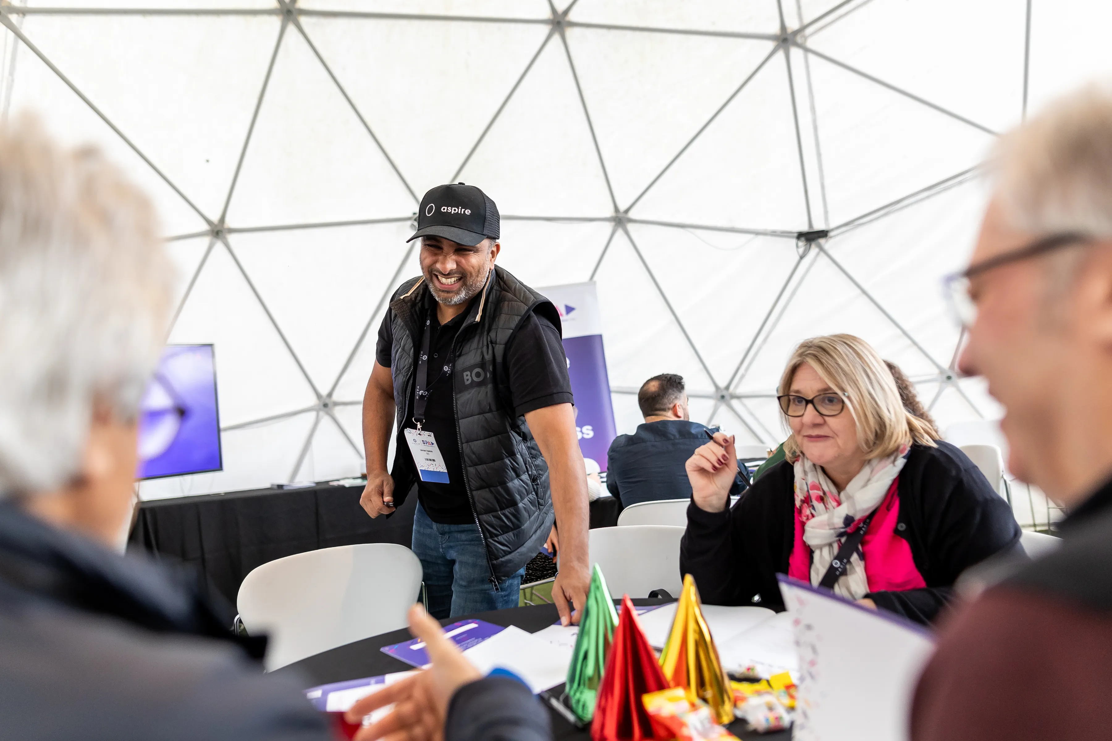 Group of adults interacting at a table with colorful party hats inside a geodesic dome tent.
