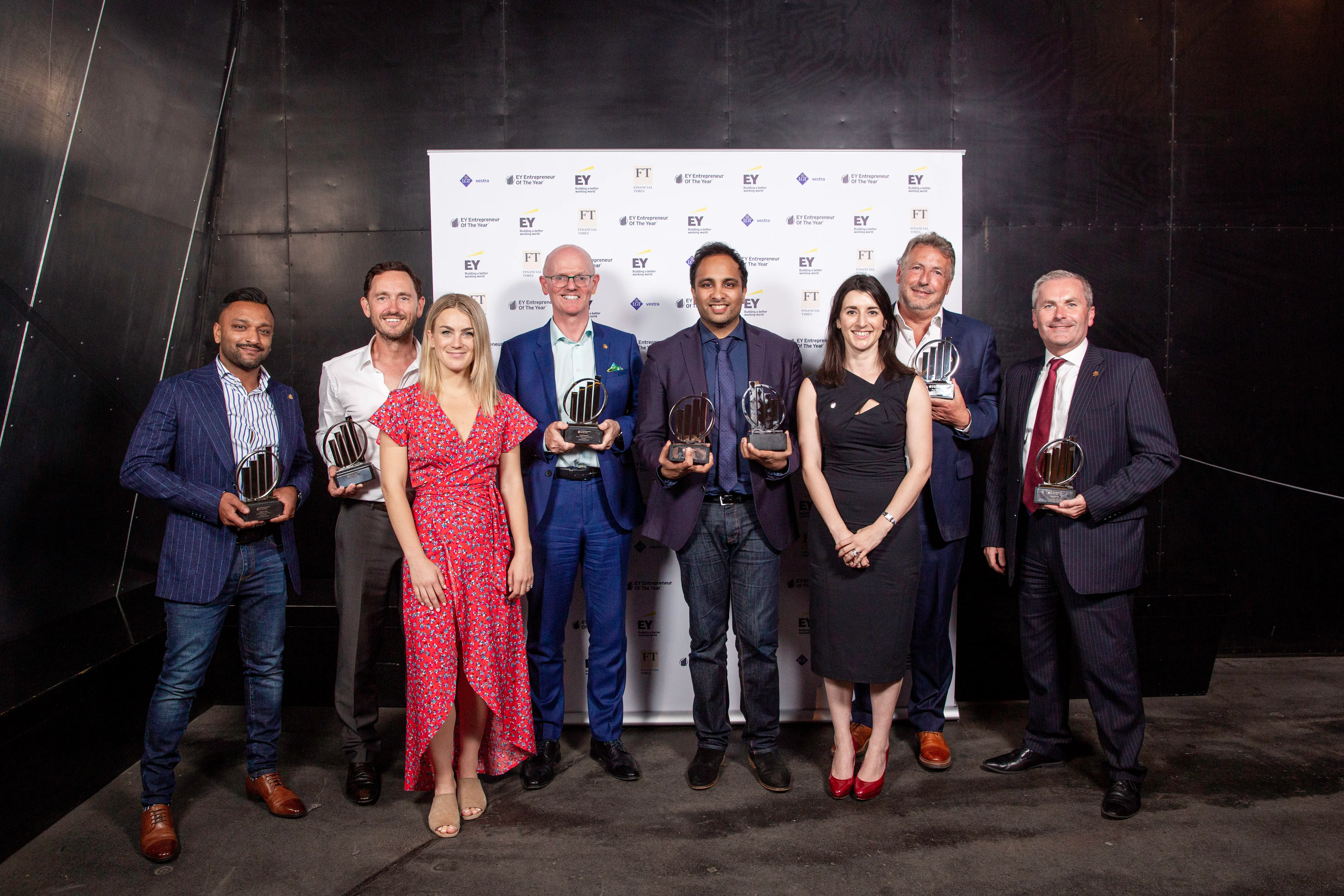 Eight award winners standing in front of a branded backdrop holding trophies and smiling.
