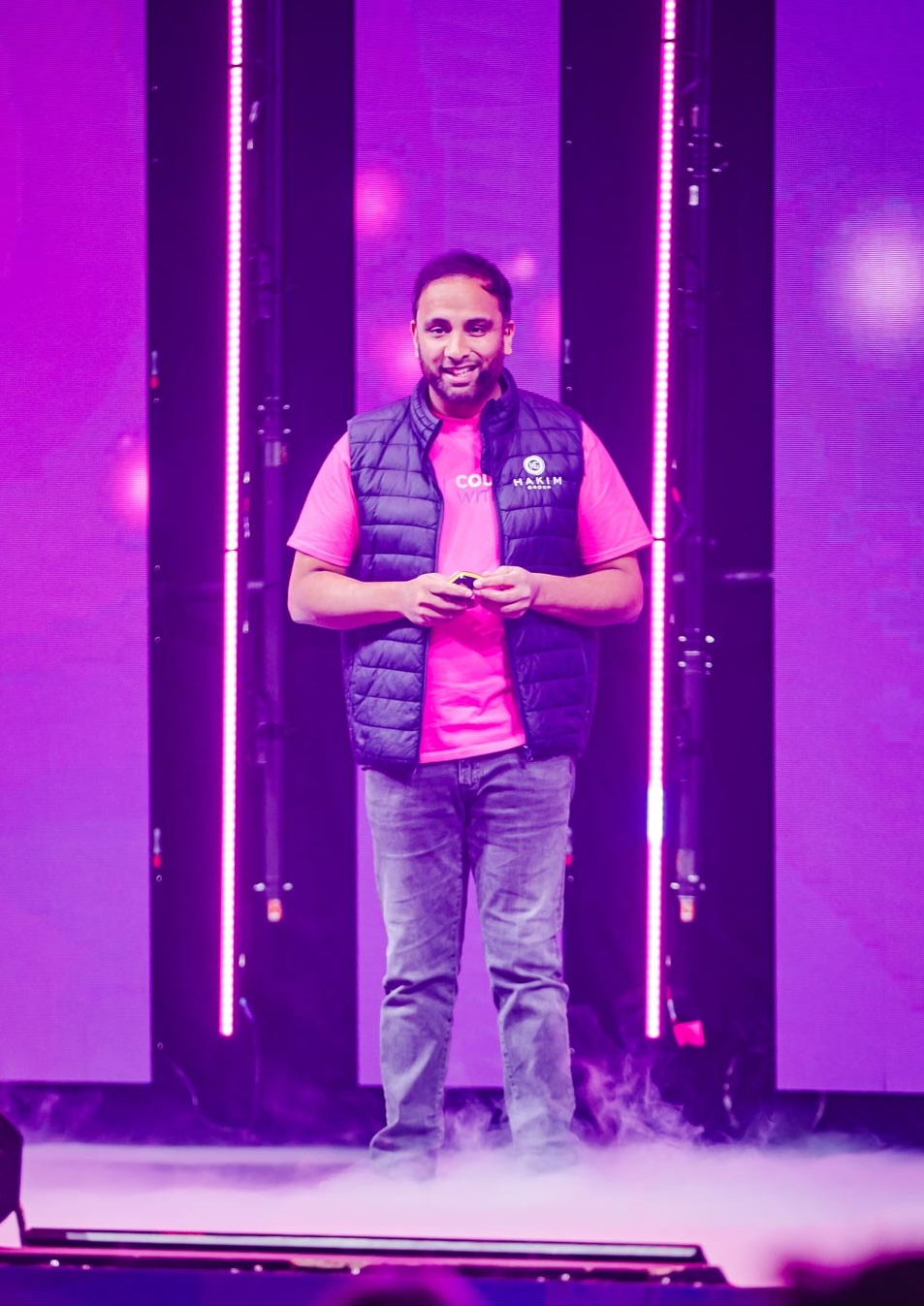 Man smiling and standing on stage with purple lights and fog, wearing a pink shirt and black vest.