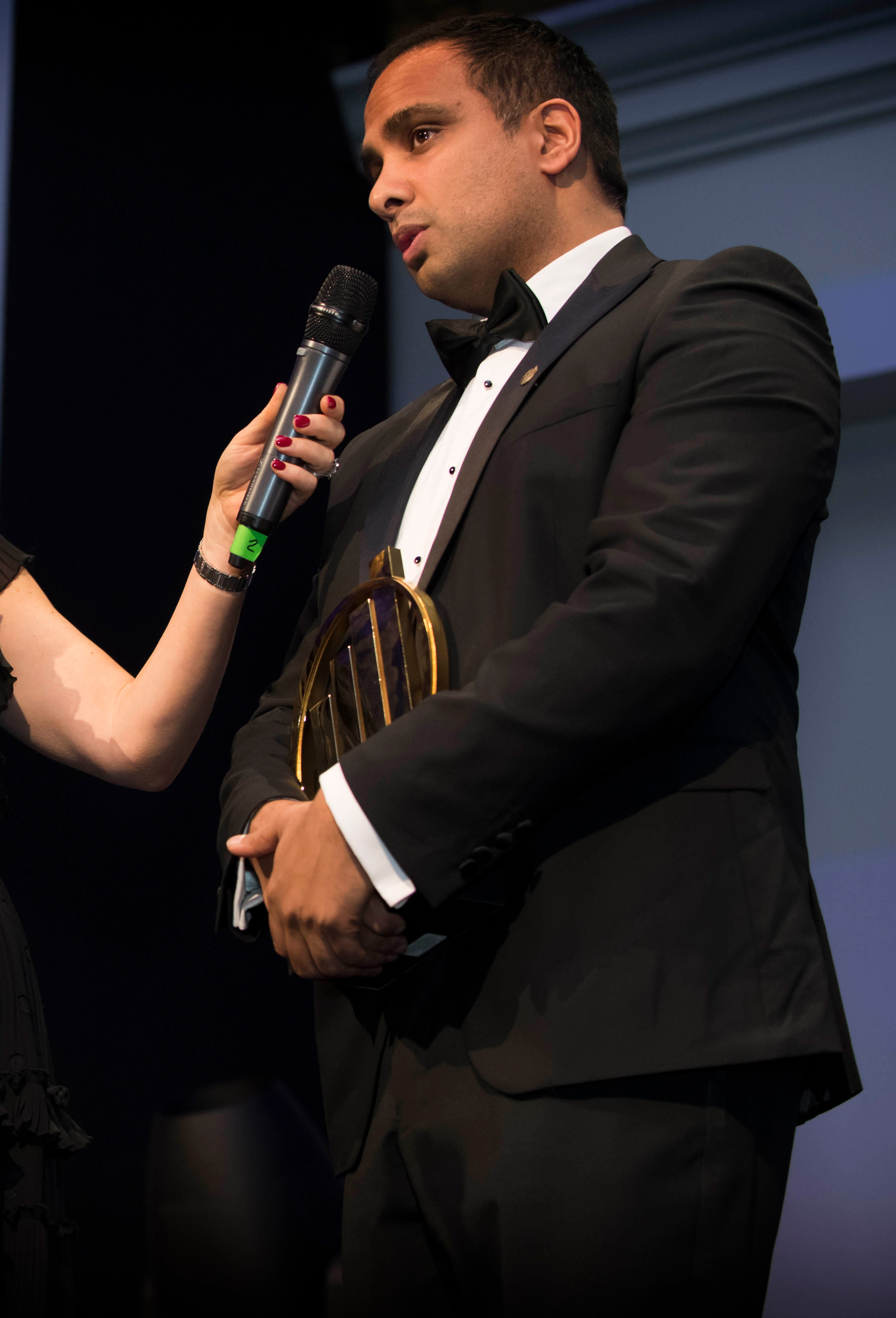 Man in black tuxedo holding an award and speaking into a microphone held by a woman.