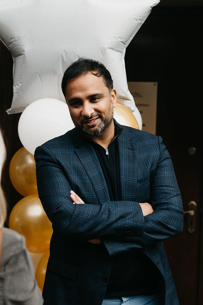 Man with folded arms smiling, standing in front of gold, white, and star-shaped balloons.