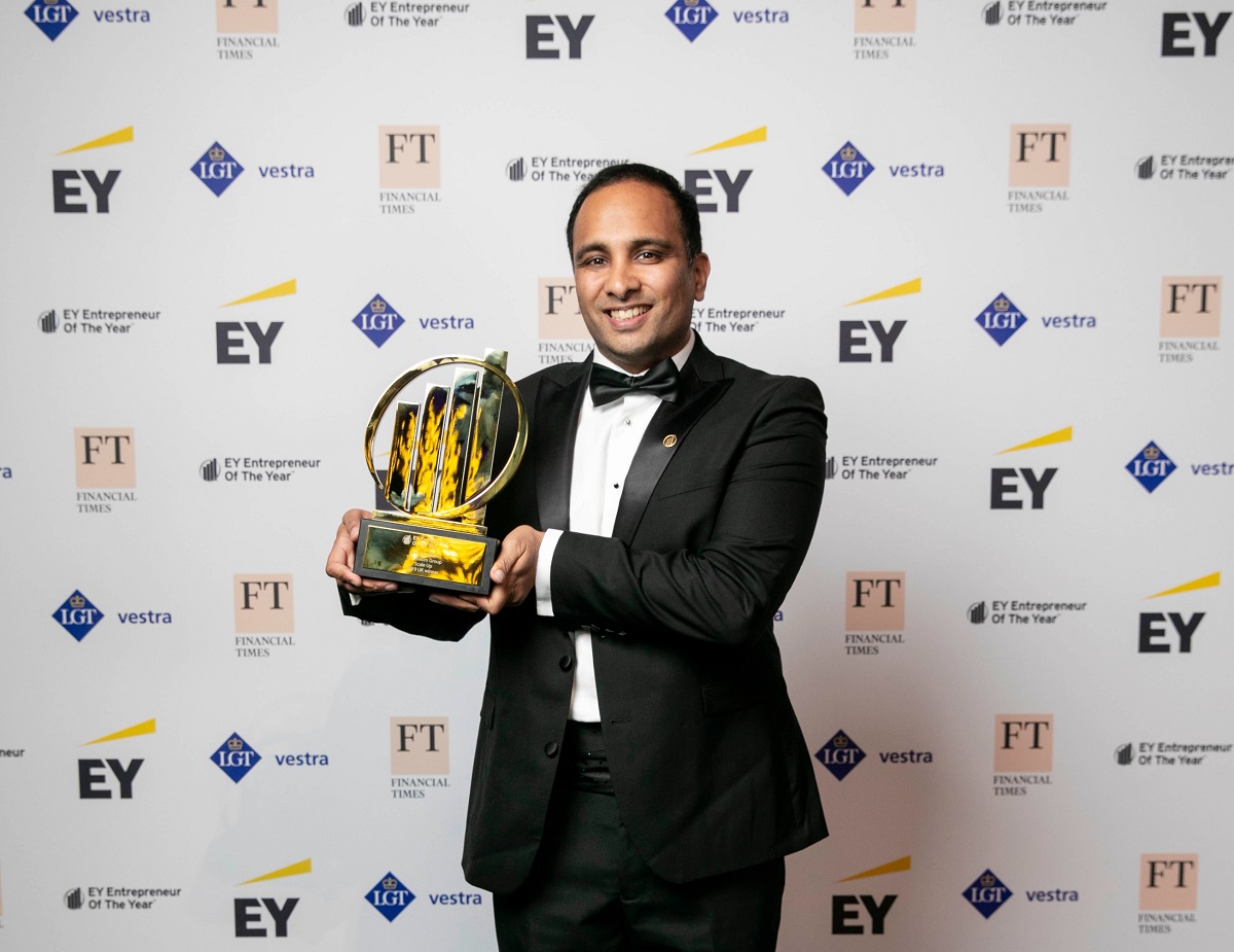 Man in tuxedo holding a gold award trophy smiling against a backdrop with EY and FT logos.