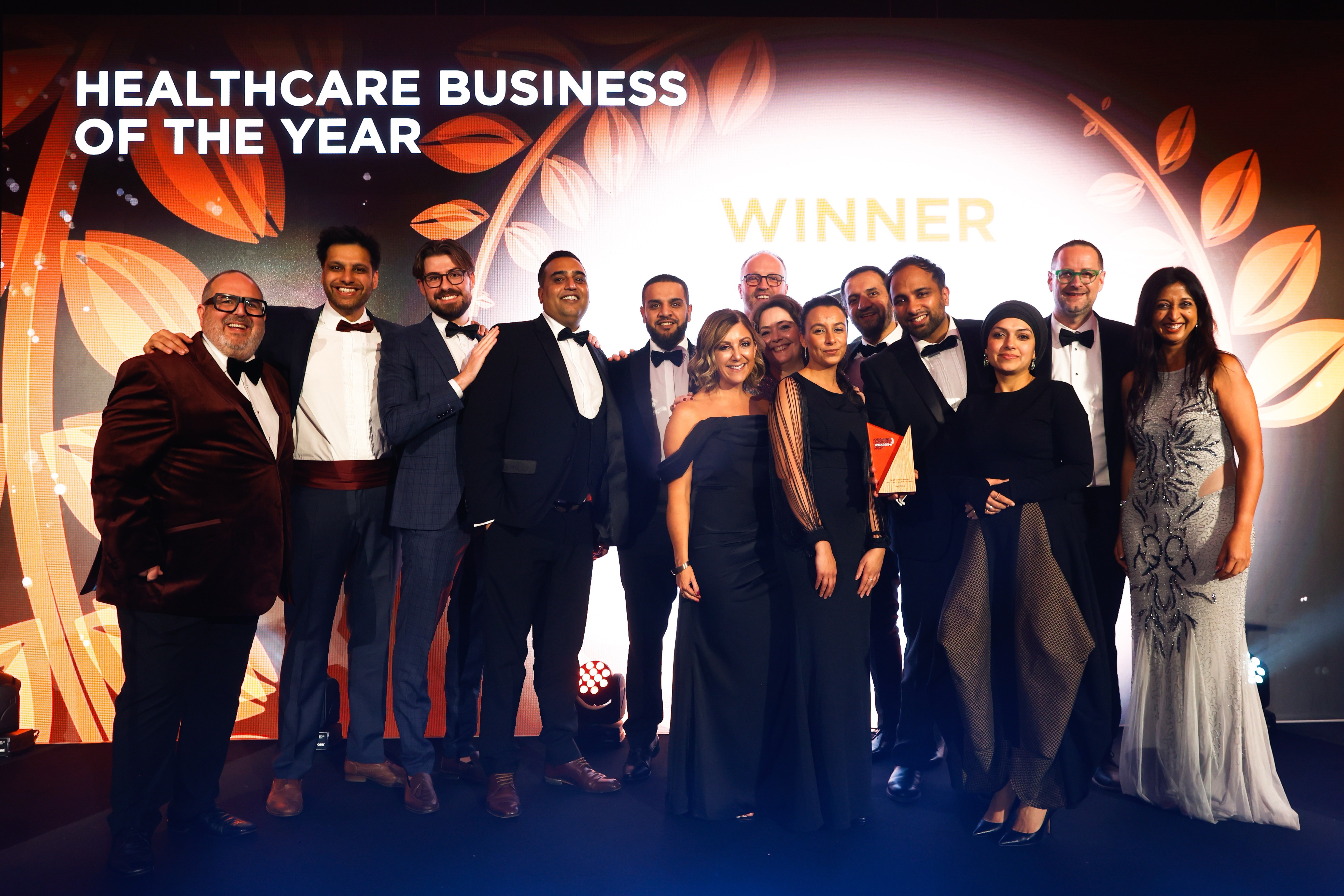 Group of people in formal attire posing with Healthcare Business of the Year award on stage.
