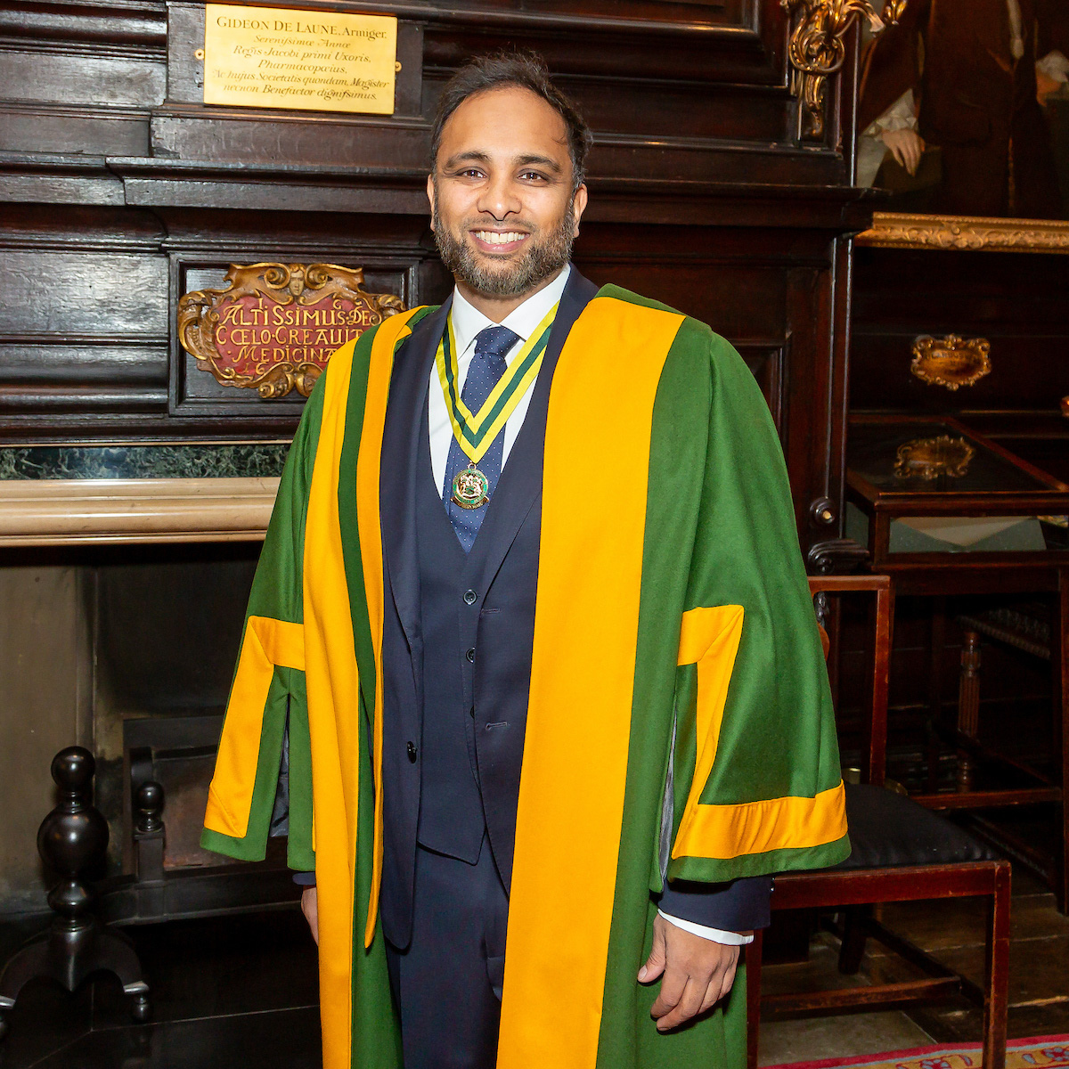 Smiling man in dark suit and green and yellow academic gown with medal around neck indoors.