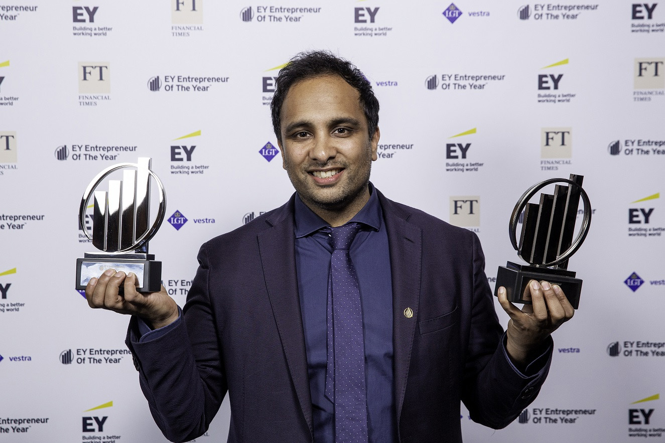 Smiling man in navy suit holding two EY Entrepreneur of the Year trophies at an award event.