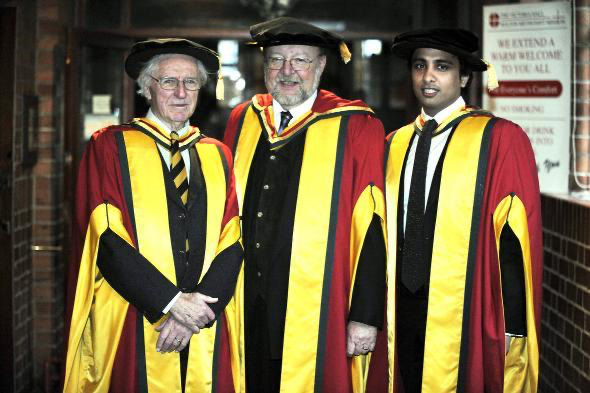 Three men in academic regalia with black caps, red and yellow robes standing indoors near brick walls.