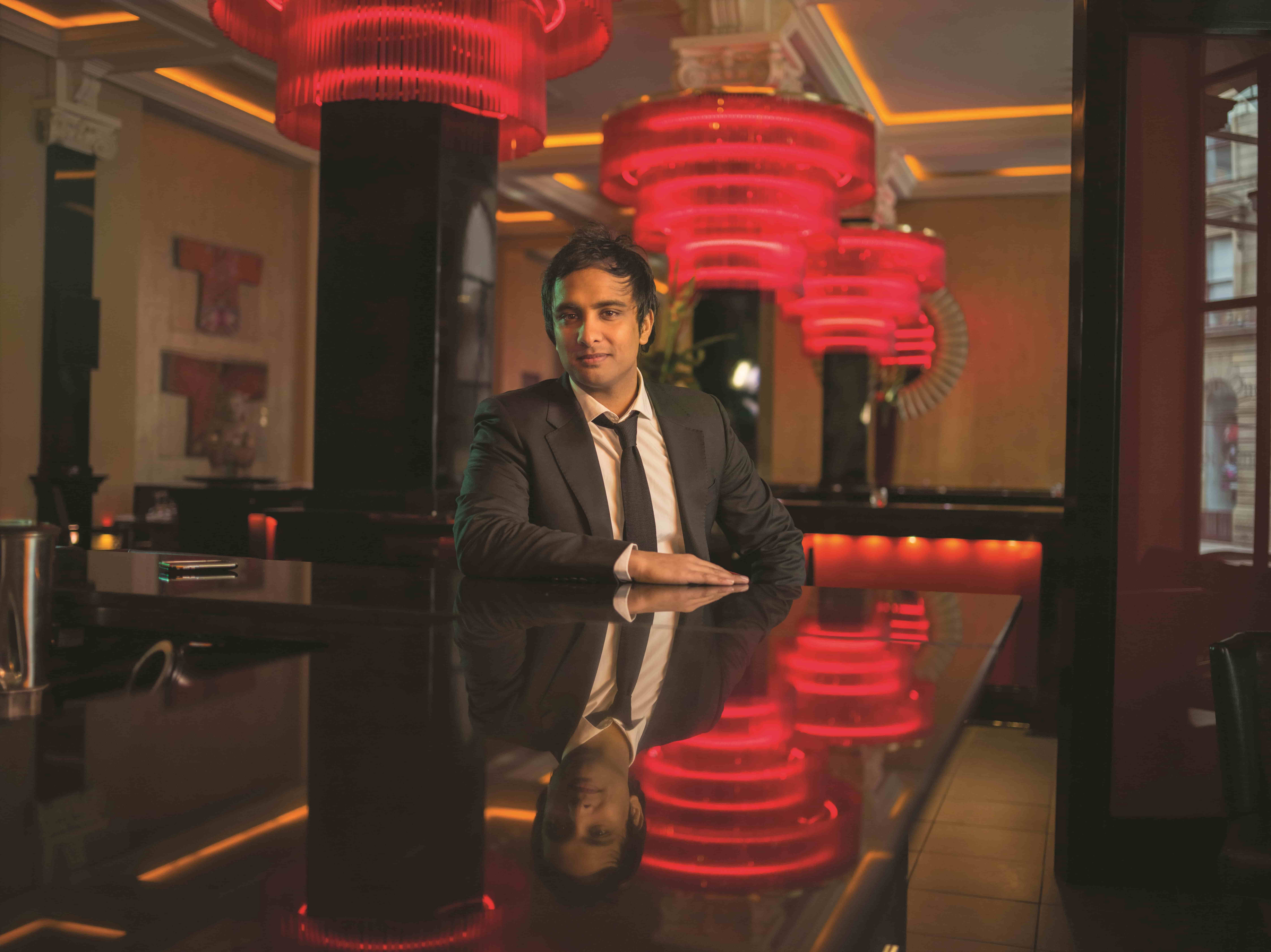 Man in black suit sitting at reflective bar counter with red lights and warm interior lighting.