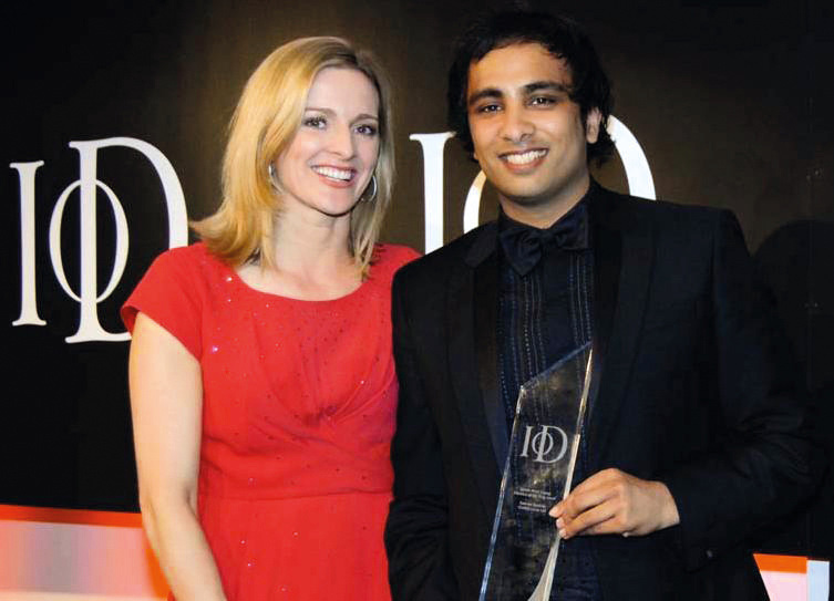 Smiling man in suit holding an award stands next to a woman in a red dress against a black backdrop.