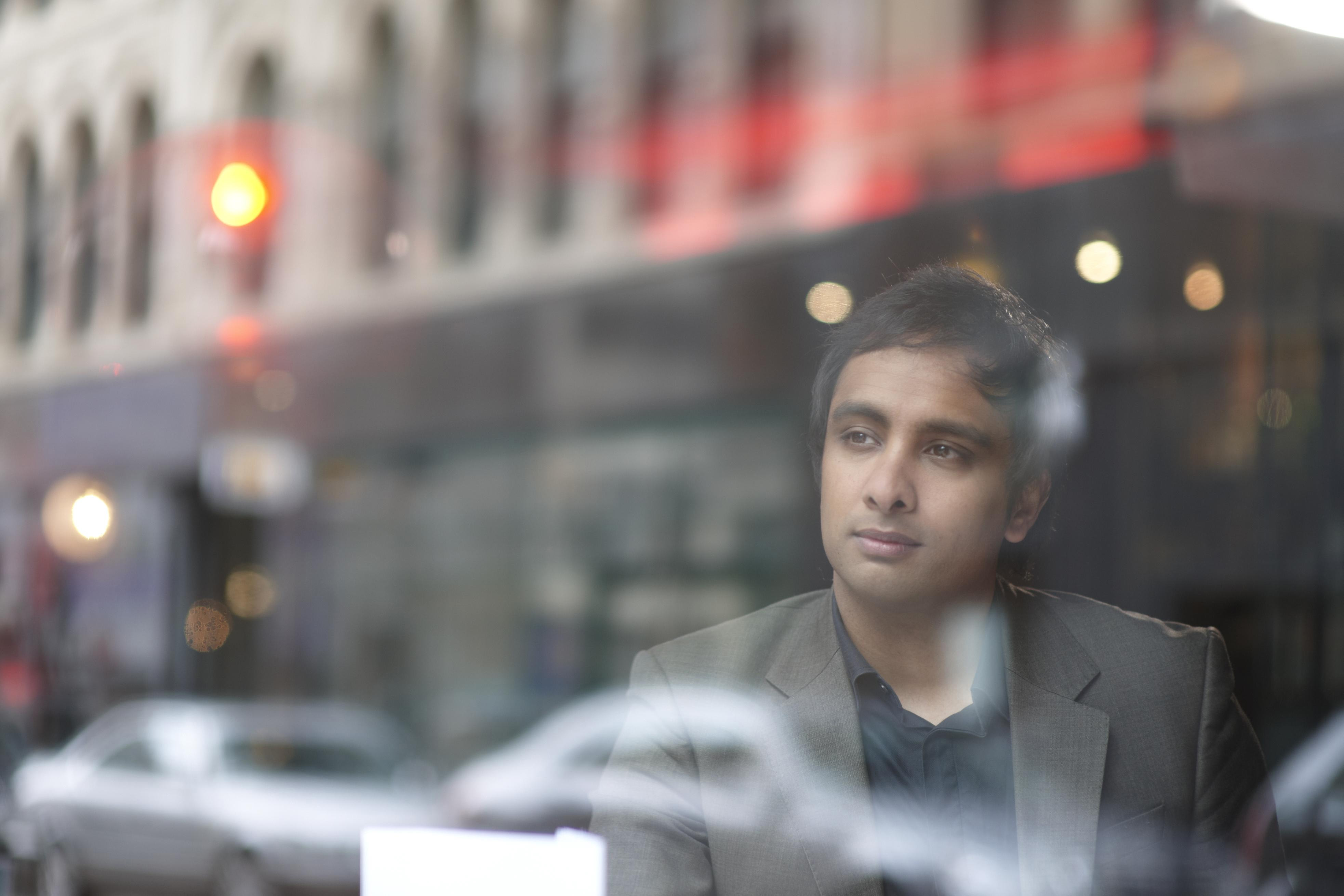 Man in a suit looking thoughtfully out a window with blurred city street reflections.