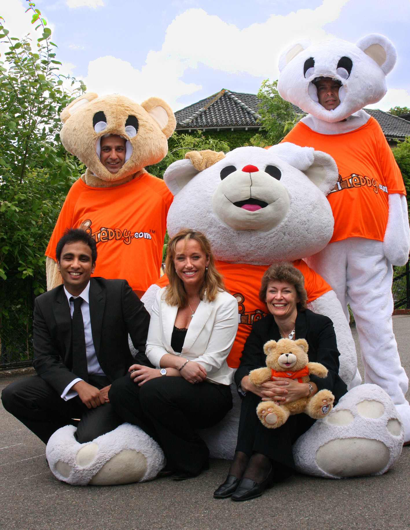 Three people in suits posing with three in orange-shirted teddy bear costumes outdoors.