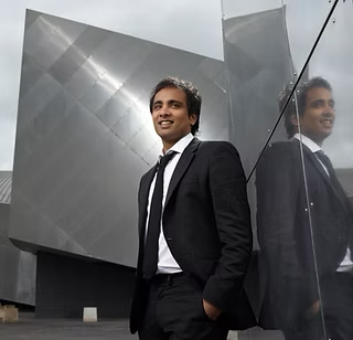 Man in a black suit standing with one hand in his pocket near a reflective glass wall and modern architecture.