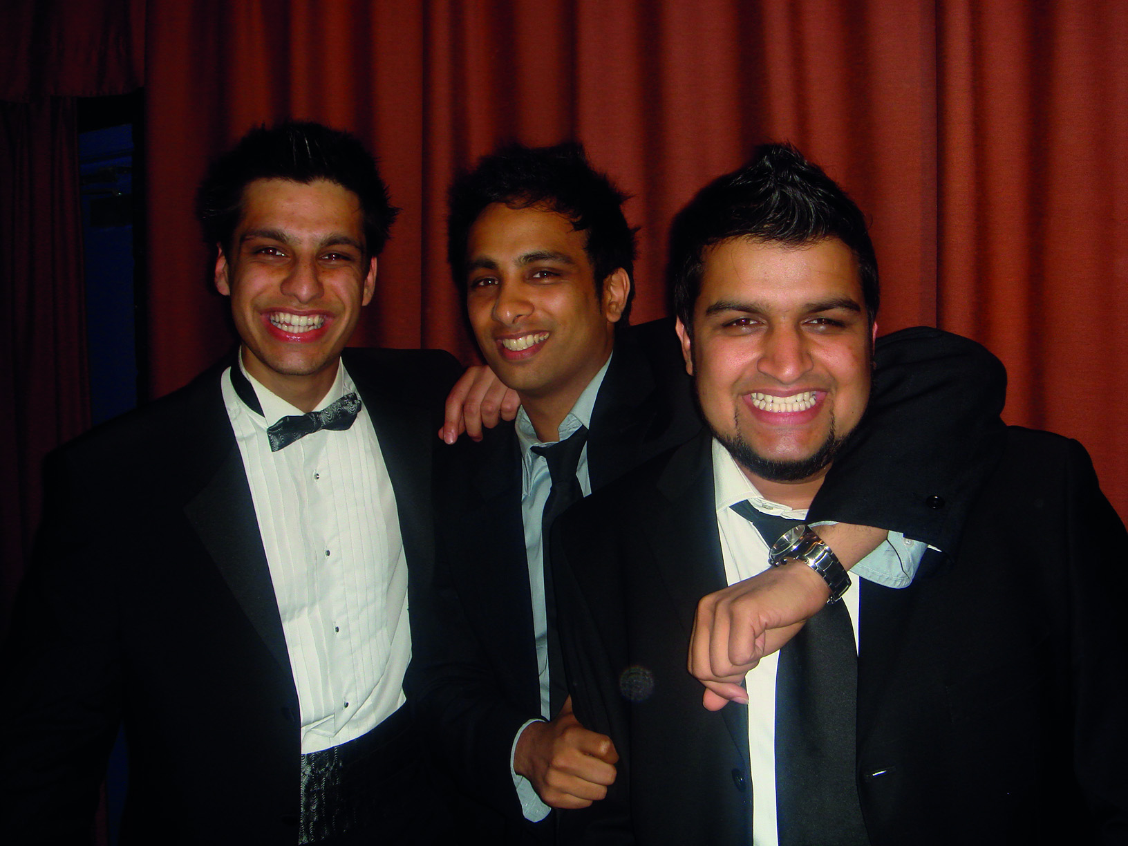 Three men in suits smiling with arms around each other against a red curtain backdrop.