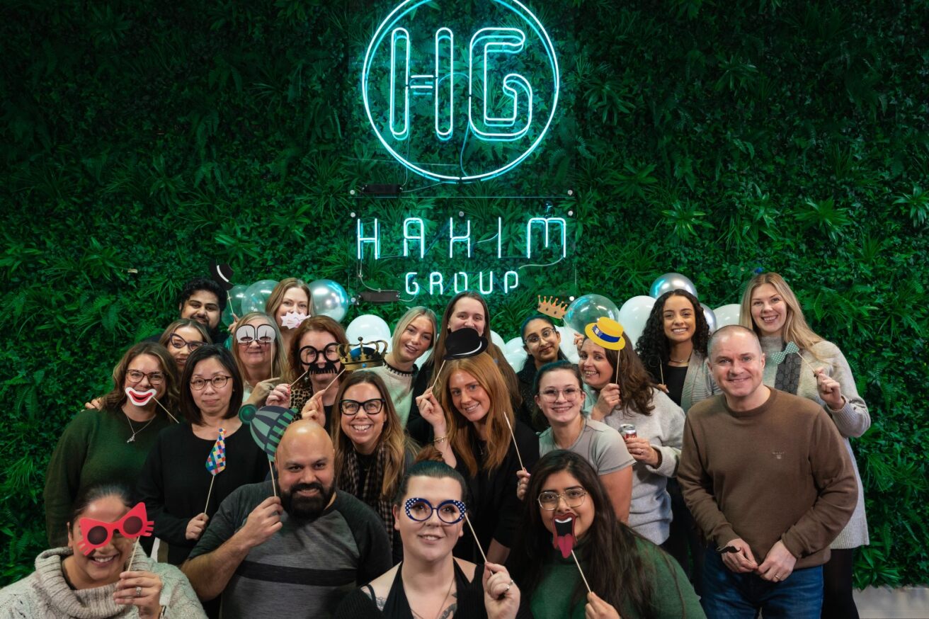 Hakim Group team celebrating together with fun props in front of the company logo wall.