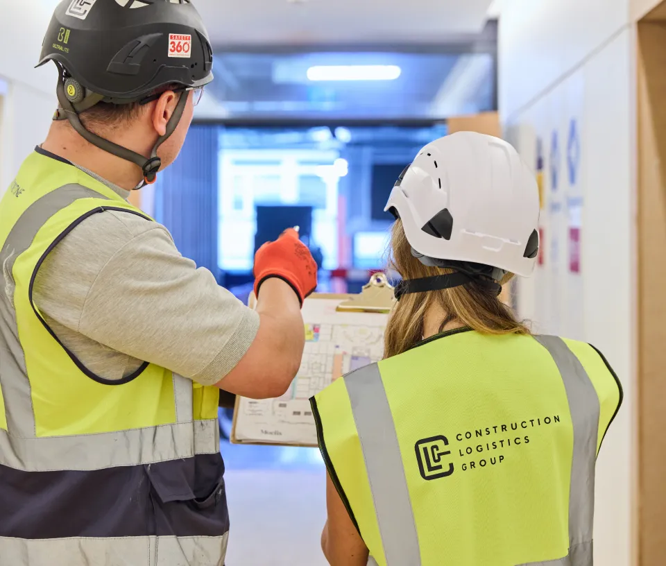 Two construction workers wearing helmets and high-visibility vests reviewing a building plan on a clipboard in a hallway.