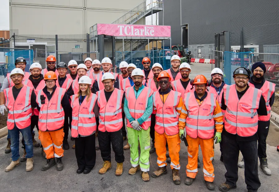 Group of construction workers wearing pink safety vests and helmets, standing together at a construction site in front of a sign that says TClarke.