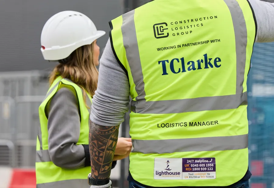 Two construction workers wearing high-visibility vests and one wearing a white helmet, with text on the vests showing Construction Logistics Group partnership with TClarke and logistics manager role.