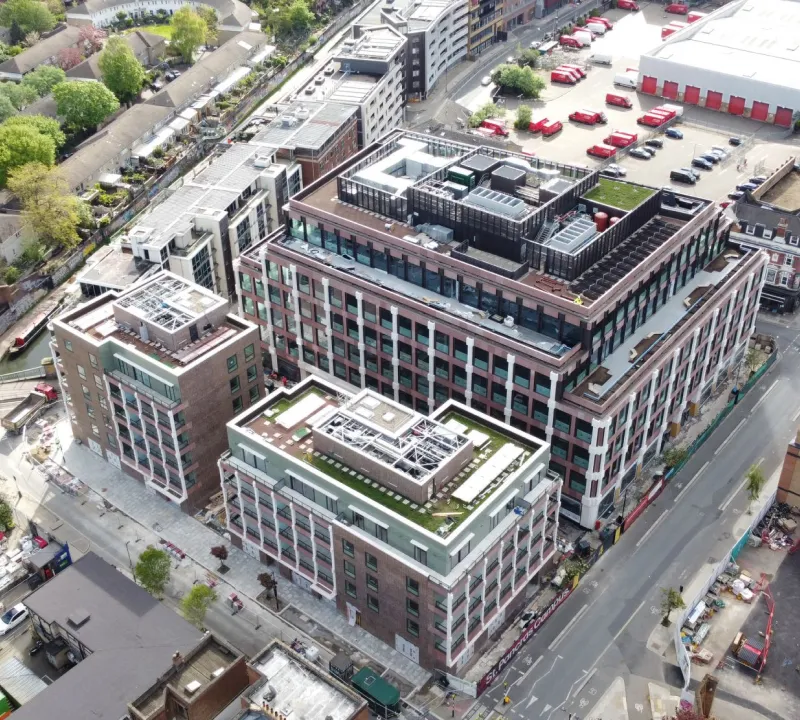 Indoor construction area with metal pipes, black support columns, a concrete pillar, and fluorescent light fixtures hanging from the ceiling.