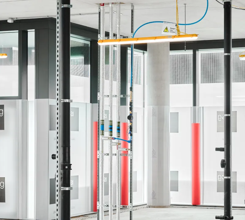 Indoor construction area with metal pipes, black support columns, a concrete pillar, and fluorescent light fixtures hanging from the ceiling.