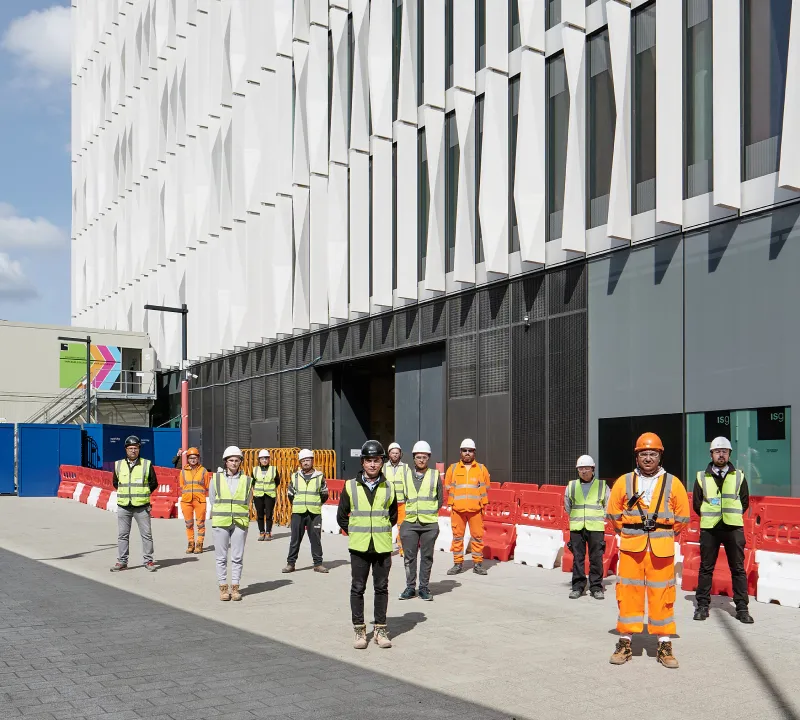 Group of construction workers wearing safety helmets and high-visibility vests standing in front of a modern building under clear sky.