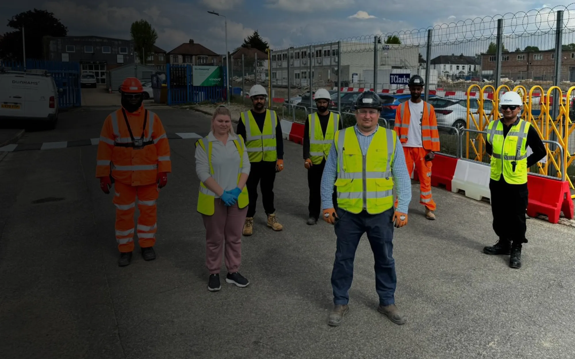 Seven Construction Logistics workers standing outdoors on a paved surface, wearing safety vests, helmets, and protective gloves, with a fenced construction site in the background.