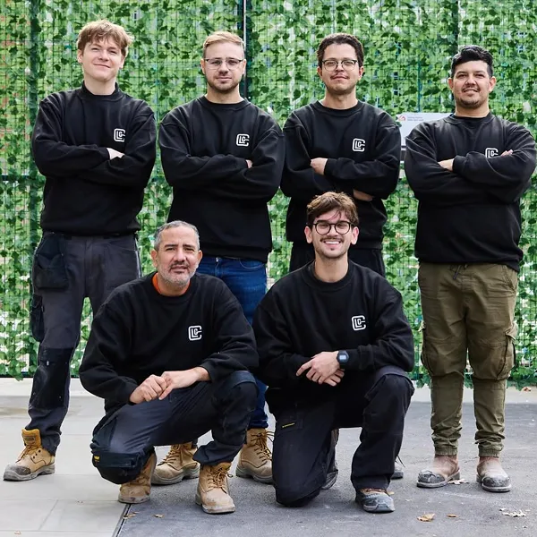 Group photo of six men wearing black sweatshirts with a white logo, standing and kneeling in front of a green leafy backdrop.