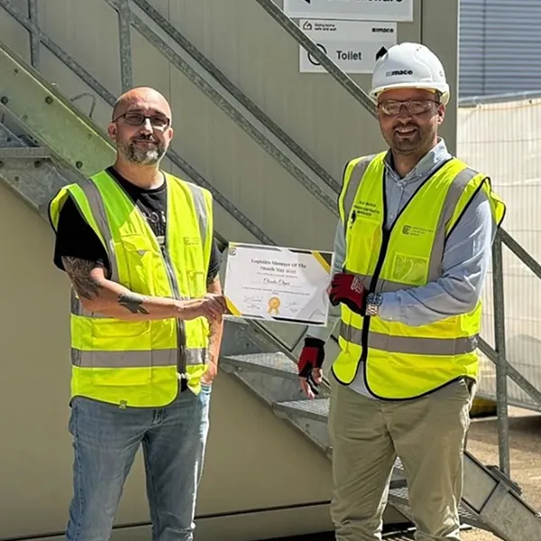 Two construction workers wearing yellow safety vests and one wearing a white hard hat, holding a certificate together.