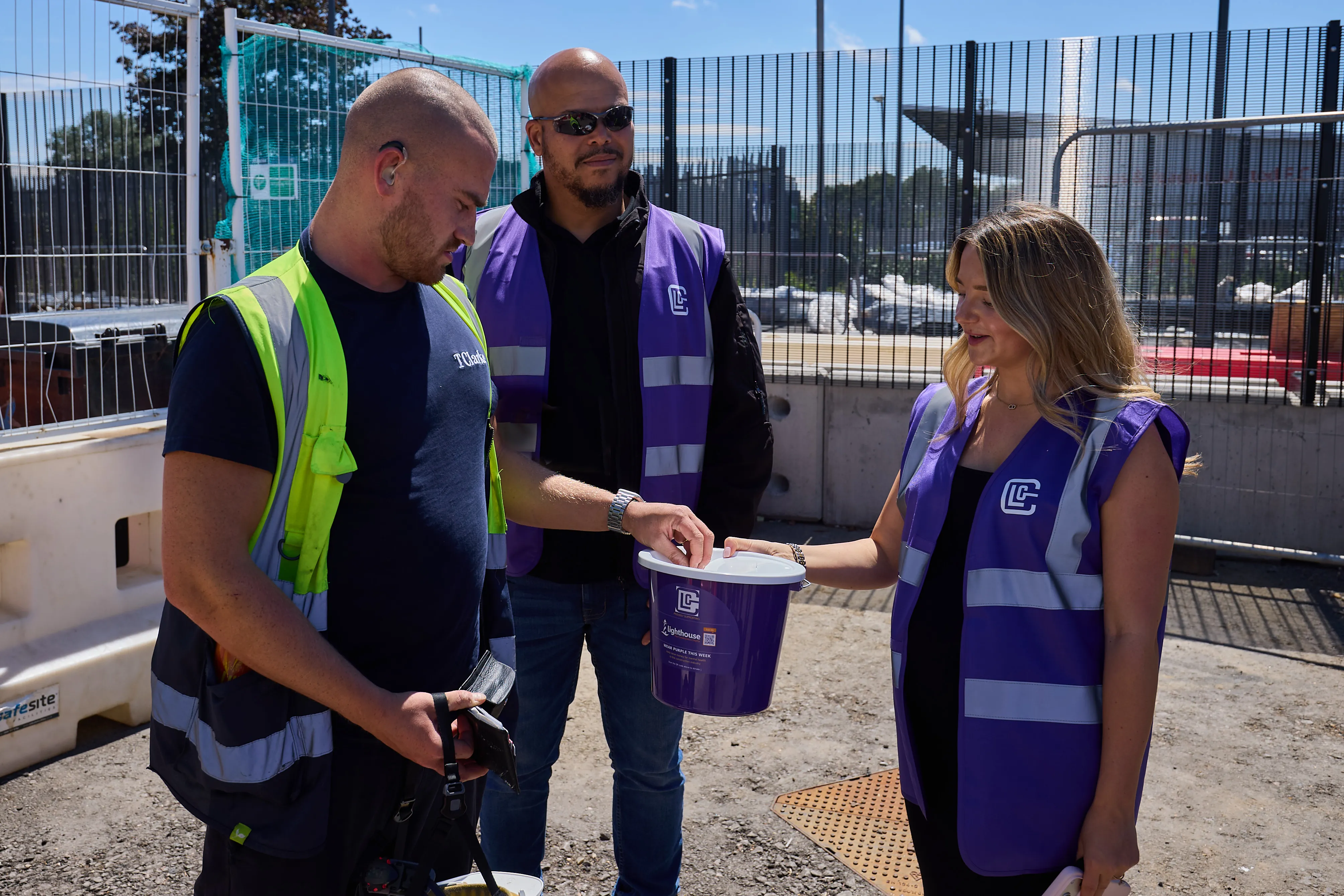 Three construction workers, two in purple vests, one in yellow vest holding a donation bucket outdoors.