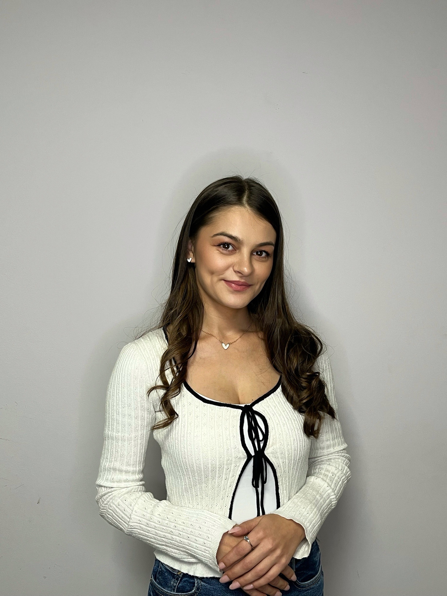 Young woman with long brown hair wearing a white sweater with black trim and blue jeans, standing against a plain light gray background.