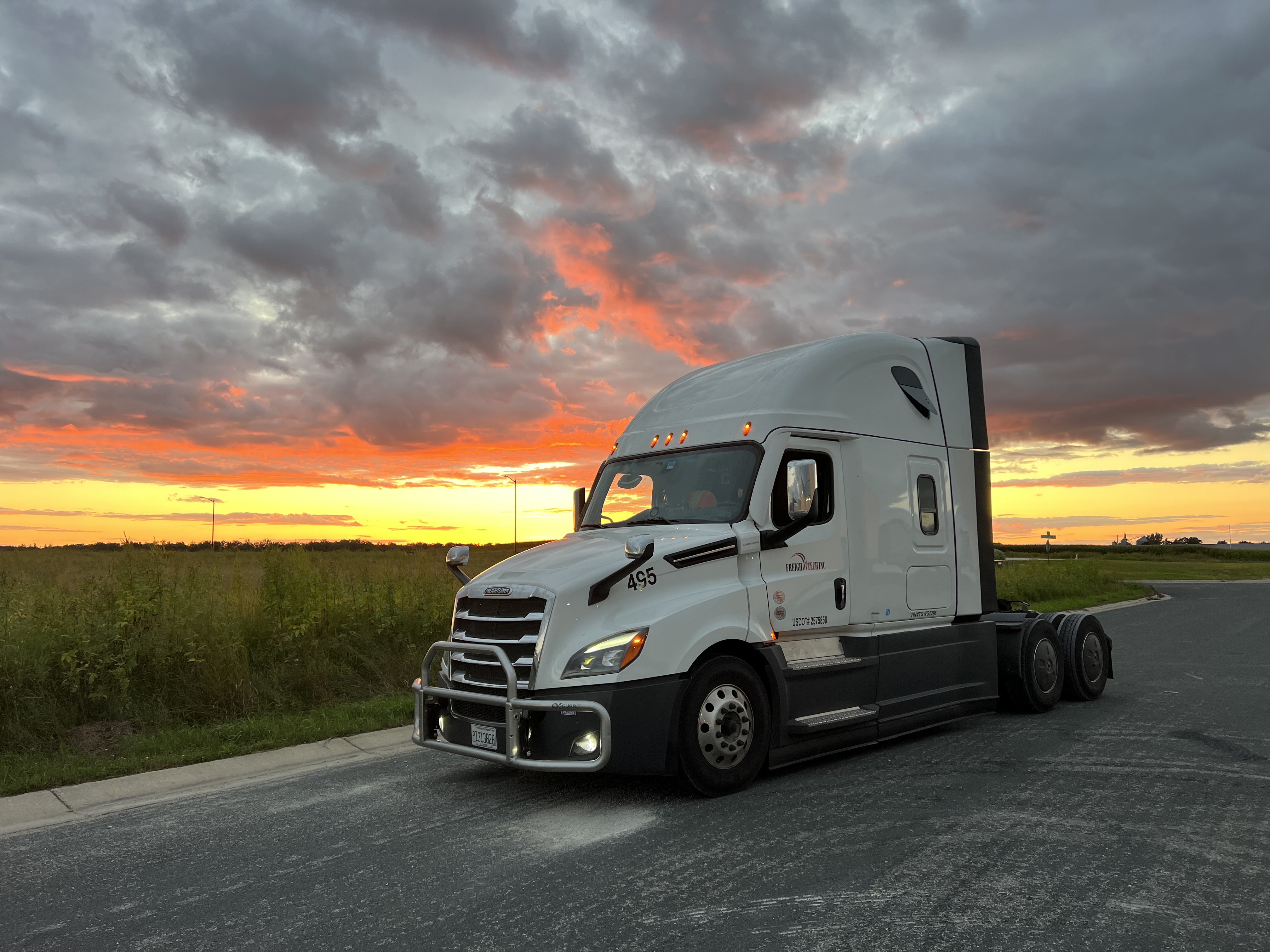 White semi-truck parked on a road beside green fields under a dramatic sunset sky with orange and gray clouds.
