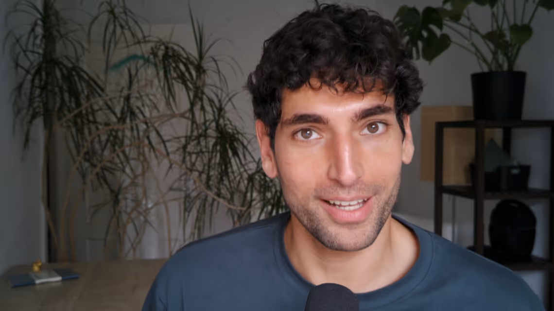 Man with dark curly hair speaking in front of a microphone indoors with plants in the background.