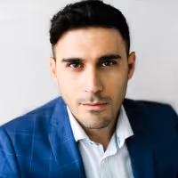 Portrait of a man with dark hair wearing a blue plaid blazer and white shirt against a plain light background.