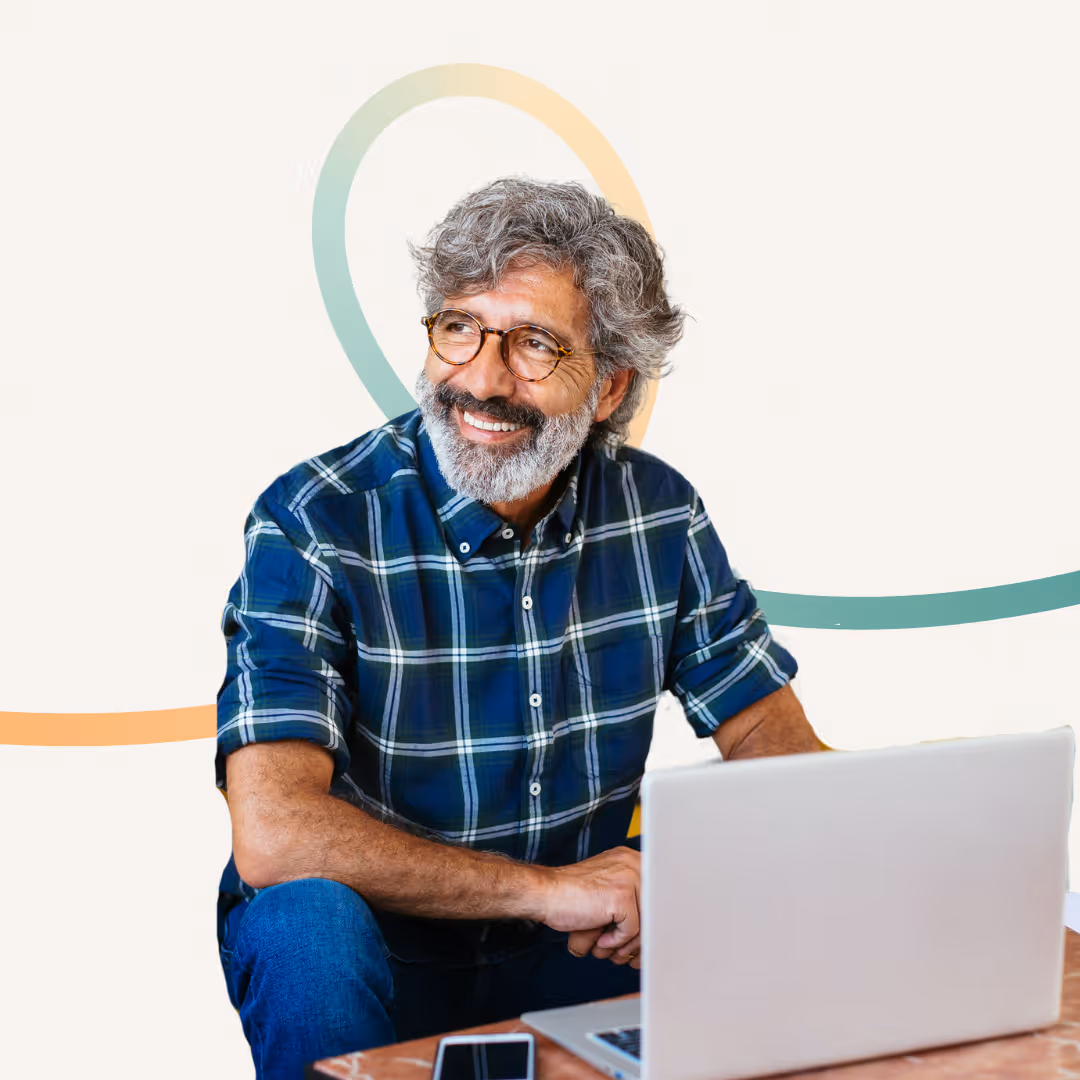 Smiling middle-aged man with gray hair and beard wearing glasses and a blue plaid shirt working on a laptop at a table.