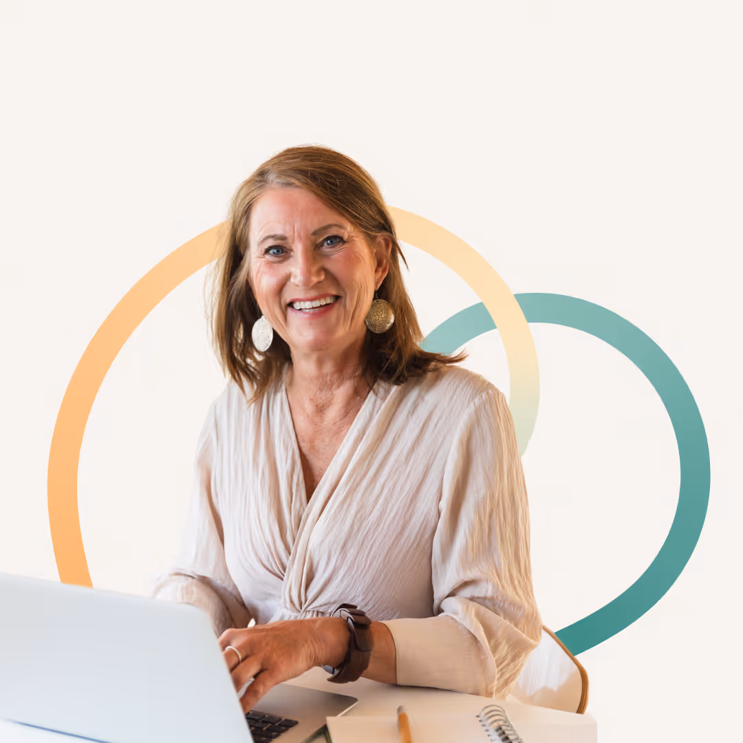 Smiling mature woman with brown hair sitting at a desk working on a laptop with a notebook and pencil nearby.