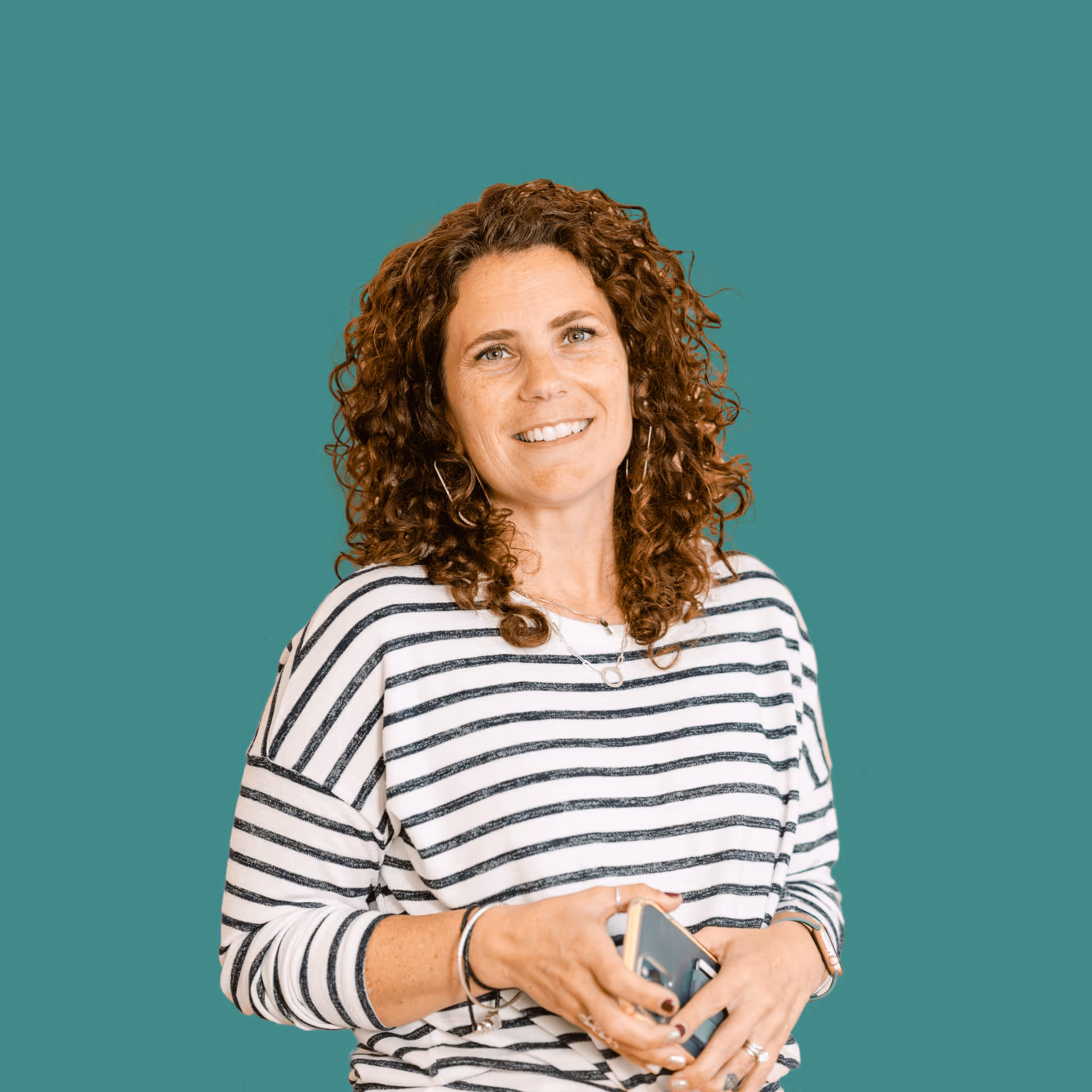 Smiling woman with curly hair wearing a striped shirt holding a smartphone against a teal background.